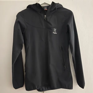 Svart vindjacka från Haglöfs - Säljer en svart vindjacka från Haglöfs med huva och dragkedja framtill. Jackan har långa ärmar och två fickor med dragkedjor. Perfekt för blåsiga dagar.