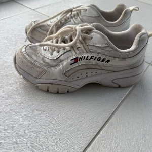 Vita sneakers från Tommy Hilfiger - Säljer ett par vita sneakers från Tommy Hilfiger med snörning och märkets logga på sidan. Skorna har en robust sula och är perfekta för en casual look. Används fåtal gånger så i fint skick. Skorna saknar däremot sulor som ni kan se på bilden men det finns att köpa sulor i de flesta skobutikerna. Skorna har reflex både fram och bak vilket är perfekt till mörkret.