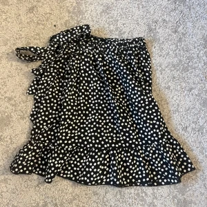 Svart blommig kjol med volang XS - Charmig svart kjol med vita blommor och volangdetaljer. Kjolen har en omlott-design med knytband i midjan för en justerbar passform. Perfekt för en somrig look! Från SHEIN i storlek XS använt en eller två gånger.