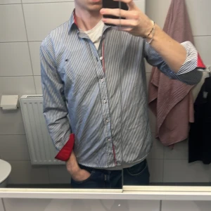 Randig Tommy hilfiger  skjorta  - Snygg randig skjorta med röda detaljer på insidan av kragen och ärmsluten. Skjortan har långa ärmar och knappar framtill. Perfekt för en stilren look.