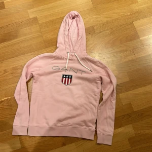 Rosa hoodie från GANT - Säljer en snygg rosa hoodie från GANT med deras ikoniska logga och ett broderat emblem på bröstet. Tröjan har en justerbar huva med dragsko och långa ärmar. Perfekt för en avslappnad stil. 💖