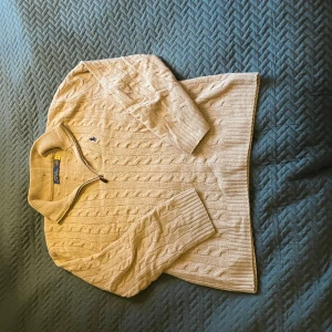 Beige kabelstickad tröja från Ralph Lauren - Säljer en stilren beige kabelstickad tröja från Ralph Lauren med dragkedja och ribbad krage. Tröjan har långa ärmar och en liten broderad logga på bröstet. Perfekt för en klassisk och avslappnad look.