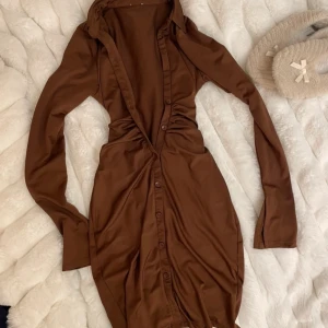 Brun klänning med knappar - Snygg brun klänning med knappar framtill och långärmad design. Klänningen har en figurnära passform och är perfekt för höst och vinter. Tillverkad i ett mjukt och bekvämt material.