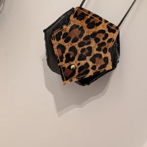 Svart axelväska med leoparddetaljer - Snygg svart axelväska i skinnimitation med krokodilmönster och leopardmönstrad ficka framtill. Väskan har en smal axelrem och stängs med en knapp. Två väskor i setet! Handsydda 