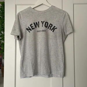 Grå t-shirt med New York-tryck från H&M - Säljer en grå t-shirt från H&M med svart text 'New York East Coast' på framsidan. T-shirten har en klassisk rund hals och korta ärmar. Perfekt för en avslappnad stil. 🗽