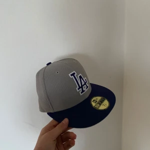 Grå och blå keps från New Era - Snygg grå och blå keps från New Era med LA-logga framtill. Perfekt för sportiga tillfällen eller som en stilren accessoar. Kepsen har en platt skärm och är en del av 59FIFTY-serien. Aldrig använd