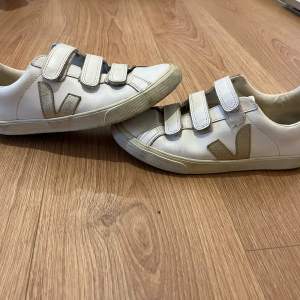 Säljer ett par skitsnygga vita sneakers från Veja med kardborreband och guldiga detaljer. Skorna har en rund tå och är tillverkade i skinn. Detaljerna är i guld. Perfekta vår och sommarskor. Har tecken på användning men fortfarande i gott skick! Fråga för fler bilder o pris kan diskuteras!!❤️