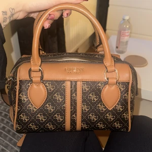 Brun och svart handväska från Guess - Säljer en snygg brun och svart handväska från Guess med deras ikoniska logotypmönster. Väskan har dubbla handtag och en avtagbar axelrem. Den är utrustad med en dragkedja och har en liten rund väska fäst vid axelremmen. Perfekt för att bära dina nödvändigheter med stil.