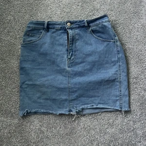 Jeanskjol i denim - Säljer en snygg jeanskjol i blå denim med en klassisk femficksdesign. Kjolen har en rå kant nertill och stängs med knapp och dragkedja framtill. Perfekt för en avslappnad stil.