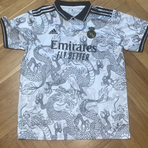 Vit fotbollströja med drakmönster - Snygg vit fotbollströja från Adidas med ett unikt drakmönster i grått. Tröjan har korta ärmar och en klassisk krage med svarta detaljer. Perfekt för fotbollsfans som vill sticka ut på planen eller läktaren.