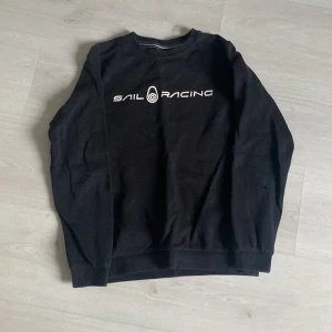 Svart tröja från Sail Racing - Säljer en svart tröja från Sail Racing med vit logotyp på bröstet. Tröjan har en klassisk design med rund hals och långa ärmar. Perfekt för en stilren och sportig look. Och det finns en liten skada på armbågen men ej synlig när man har på den.