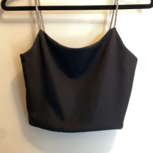 Svart tubtopp med glittriga axelband - Snygg svart tubtopp med glittriga axelband som ger en festlig touch. Perfekt för en utekväll eller speciella tillfällen. Enkel och stilren design som passar till många olika outfits. Den är i nyskick från New Yorker i storlek XXS
