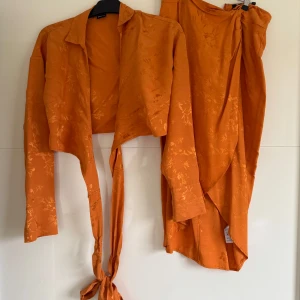 Orange omlottkjol - Snygg orange omlottkjol med subtilt blommönster. Kjolen har en knytning i midjan och är perfekt för en färgglad look. 2 delar set. 