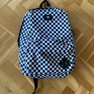 Svartvit rutig ryggsäck från Vans - Snygg svartvit rutig ryggsäck från Vans med klassiskt schackmönster. Ryggsäcken har justerbara axelremmar och en praktisk dragkedja framtill. Perfekt för skolan eller vardagsbruk. Inga fläckar och inget trasigt