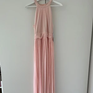 Ljusrosa långklänning  - Elegant ljusrosa långklänning köpt från Bubbleroom, med ett vackert fall och knytning i nacken. Klänningen har en ärmlös design och ett plisserat kjolparti som ger en romantisk känsla. Perfekt för tillfällen som bal. 