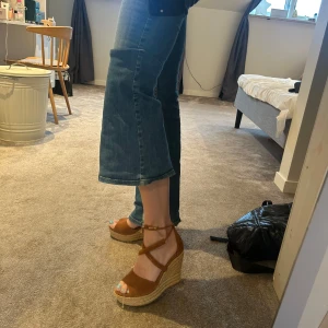 Bruna kilklackar från UGG - Snygga bruna kilklackar från UGG i mocka med knytning runt ankeln. Perfekta för sommarens alla tillfällen. Klackarna har en flätad detalj som ger en bohemisk känsla.