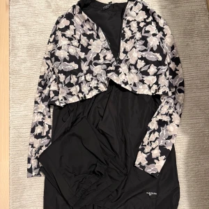 Svart klänning med blommigt mönster från Andiata - Elegant svart klänning från Andiata med ett vackert blommigt mönster i ljusa toner. Klänningen har långa ärmar och en omlott-design framtill. Perfekt för en stilfull look.