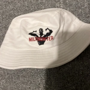 Vit bucket hat med tryck - Snygg vit bucket hat med ett tryck av en siluett och texten 'MILFHUNTER' i rött. Perfekt för en avslappnad stil med en touch av humor. Hatten har en klassisk form och är tillverkad i ett lätt material.