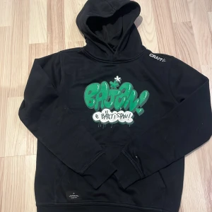 Svart Bajen hoodie från Craft - Snygg svart hoodie från Craft med grön och vit graffiti-design på framsidan. Tröjan har en klassisk känguruficka och en bekväm huva. Perfekt för en avslappnad stil.