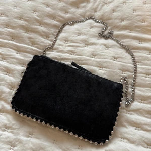 Svart väska med kedja och pärldetaljer - Snygg svart väska med en elegant kedja och pärldetaljer längs kanterna. Perfekt för att ge en touch av glamour till din outfit. Väskan har en dragkedja för säker förvaring.