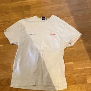 Axel arigato tshirt - Säljer en beige t-shirt från Axel Arigato med ett coolt hjärtmotiv på baksidan. T-shirten har korta ärmar och en avslappnad passform. Perfekt för en casual look! I princip ny