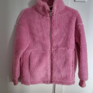 Rosa teddyjacka med dragkedja - Mysig rosa teddyjacka med dragkedja framtill. Perfekt för kyliga dagar med sin fluffiga och varma design. Jackan har långa ärmar och en hög krage för extra värme.