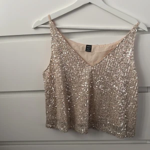 Glittrig beige paljettopp från H&M - Toppen är ärmlös med en v-ringad design och täckt av paljetter som ger en festlig look. Perfekt för en utekväll eller festliga tillfällen. ✨     