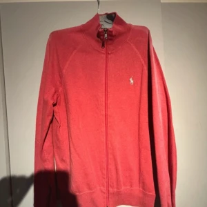 Röd tröja med dragkedja från Polo Ralph Lauren - Säljer en stilren röd tröja med dragkedja från Polo Ralph Lauren. Tröjan har en liten broderad logotyp på bröstet och långa ärmar. Perfekt för en avslappnad och snygg look. Liten missfärgning och ett litet hål