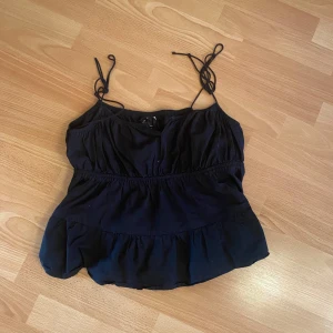 Svart linnetopp från H&M Divided - Säljer en svart linnetopp från H&M Divided med knytbara axelband och volangdetaljer. Perfekt nu inför sommaren för söta outfits. Axelbanden är justerbara vilket passar alla! Storlek 44 men är ganska liten i storleken så passar till mindre storlekar!!