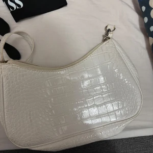 Vit axelväska med krokodilmönster - Snygg vit axelväska med krokodilmönster och dragkedja. Perfekt för att ge en stilren touch till din outfit. Väskan har en justerbar axelrem och är tillverkad i syntetmaterial.