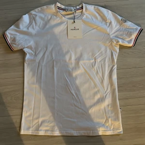 Vit t-shirt från Moncler - Säljer en stilren vit t-shirt från Moncler med korta ärmar och snygga randiga detaljer i rött och blått vid ärmslut och krage. Perfekt för en casual look. Moncler-logga på ärmen ger en exklusiv touch.