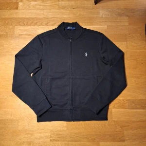 Svart tröja med dragkedja från Polo Ralph Lauren - Säljer en stilren svart tröja från Polo Ralph Lauren med dragkedja och broderad logotyp på bröstet. Tröjan har långa ärmar och en klassisk design som passar perfekt för olika tillfällen.