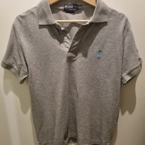 Grå pikétröja från Ralph Lauren - Grå pikétröja från Ralph Lauren med korta ärmar och en broderad logga på bröstet. Litet hål i nacken vid lappen, därav priset.