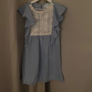 Blå klänning med spetsdetaljer från Zara - Säljer en söt blå klänning från Zara med vackra vita spetsdetaljer framtill. Klänningen har volanger längs sidorna och är ärmlös. Köpt på baravdelningen men kommer passa en xxs/xs