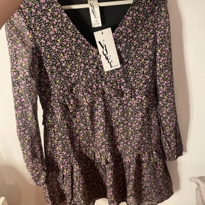 Blommig klänning från ASOS - Säljer en söt blommig volangklänning från Violet Romance i lila och svart. Köpt på asos. Klänningen har långa ärmar och en v-ringad design. Helt ny med prislapp