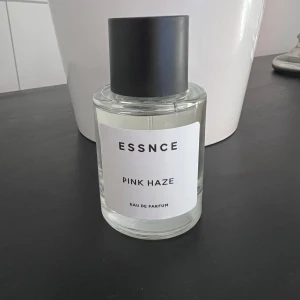 Essnce Pink Haze Eau de Parfum - Upptäck Essnce Pink Haze, en Eau de Parfum med en fräsch och fruktig doftprofil. Flaskan är elegant och minimalistisk med en svart kork och en klar glasdesign. Doften kombinerar toner av bär, apelsin och blommor, vilket ger en söt och upplyftande känsla. Perfekt för dig som älskar en livlig och energisk doft.