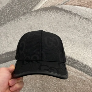 Svart keps från Gucci - Snygg svart keps från Gucci med diskret mönster av märkets logotyp. Kepsen har en justerbar rem baktill för perfekt passform. Perfekt för att ge en stilren touch till din outfit.