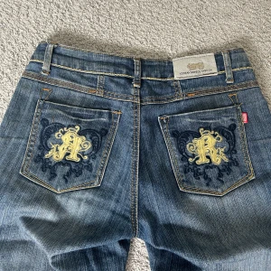 Blå jeans med broderad detalj - Coola jeans med unika broderade detaljer. Midjemåttet är 36 cm rakt över, innerbenslängden är ca 67 cm. Det fattas några pärlor på bakfikorna men man märker knappt. Dessa jeans köptes på Plick men säljs för att de är för korta på mig.