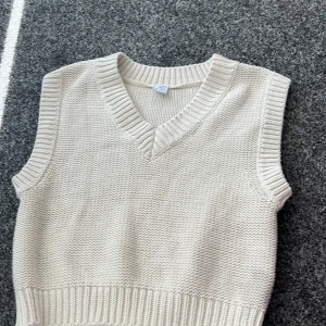 Beige stickad väst från Lindex - Säljer en stilren beige stickad väst från Lindex för barn. Västen har en v-ringad design och ribbade kanter som ger en klassisk look. Perfekt för lager-på-lager under kyligare dagar.