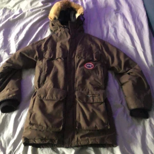 Brun parkajacka från Canada Goose - Säljer en brun parkajacka från Canada Goose med pälsdetaljer på huvan. Jackan har flera praktiska fickor och en broderad logotyp på bröstet. Perfekt för kalla dagar.