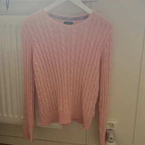 Rosa stickad tröja från Ralph Lauren - Säljer en söt rosa stickad tröja från Ralph Lauren med klassiskt flätmönster. Tröjan har långa ärmar och en rund halsringning. Perfekt för kyligare dagar. 🌸