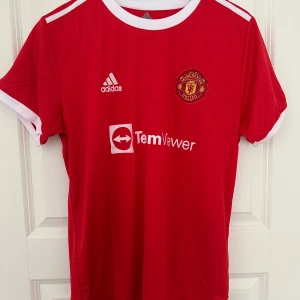 Röd Manchester United fotbollströja - Säljer en röd Manchester United fotbollströja från Adidas med korta ärmar. Tröjan har klubbens emblem på bröstet och sponsorlogga. På baksidan finns texten 'Ronaldo' och nummer 7. Perfekt för fans av laget!