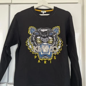 Svart tröja med tigerbroderi från Kenzo - Säljer en svart tröja från Kenzo med ett stort, färgglatt tigerbroderi på framsidan. Tröjan har långa ärmar och en rund halsringning. Perfekt för dig som vill ha en statement piece i garderoben!