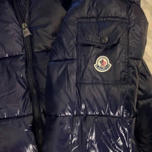 Blå dunjacka från Moncler - Säljer en snygg blå dunjacka från Moncler med glansig finish. Jackan har en dragkedja framtill och en praktisk ficka med knapp på ärmen. Perfekt för kyliga dagar. Använd fåtal gånger, säljer den pågrund av att det ej är min stil!