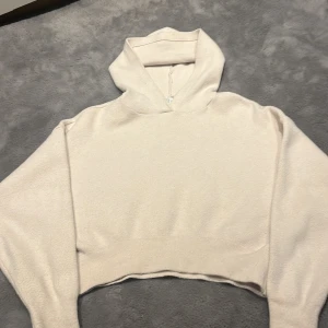Beige croppad hoodie - Säljer en stilren beige croppad hoodie med en mysig huva. Perfekt för en avslappnad look. Den har långa ärmar och en ribbad nederkant som ger en snygg passform.