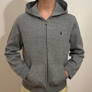Grå hoodie från Ralph Lauren - Säljer en stilren grå hoodie från Ralph Lauren med dragkedja och det klassiska logotypbroderiet på bröstet. Tröjan har långa ärmar och en praktisk huva. Perfekt för en avslappnad look!