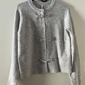 Grå kofta från Nelly - Säljer en stilren grå kofta från Nelly med dekorativa knytrosetter framtill. Koftan har långa ärmar och ribbade muddar vid ärmslut och nederkant. Perfekt för kyligare dagar.