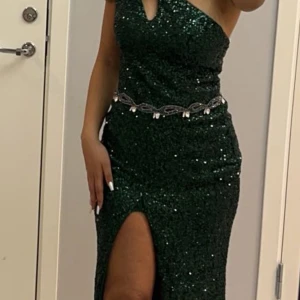 Grön glittrig klänning med slits - Snygg grön glittrig klänning med en elegant slits framtill och en öppen rygg. Klänningen har en v-ringad design och ett dekorativt bälte i midjan. Perfekt för en festlig kväll! 
