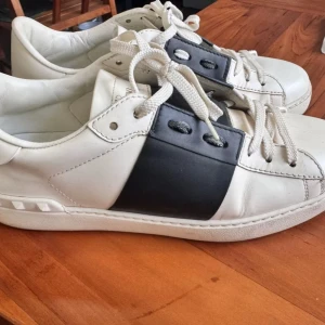 Valentino Garavani sneakers i vitt och svart - Snygga Valentino Garavani sneakers i vitt med en bred svart rem över snörningen. Skorna har en klassisk design med vita snören och en bekväm sula. Perfekta för en stilren look. Storlek 40