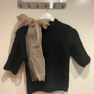 Svart tröja med beige rosett - Säljer en stilren svart tröja med en elegant beige rosett vid axeln. Tröjan har korta ärmar och är tillverkad i ett mjukt material som ger en lyxig känsla. Perfekt för att ge en touch av elegans till din outfit.